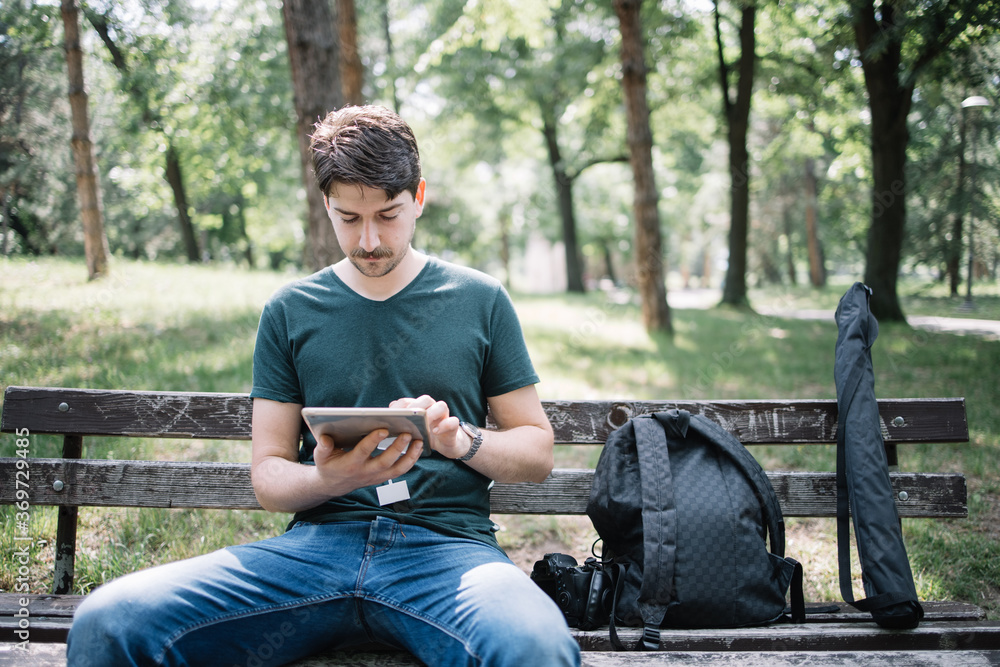 Obraz premium Young male photographer resting on wooden seat in park. Handsome man sitting on wooden bench in park and using tablet.