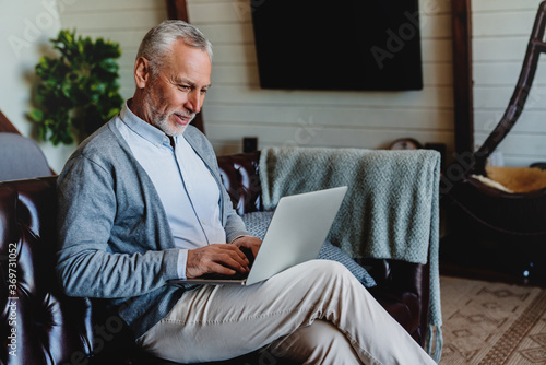 Картината върху платно Handsome old man using laptop and smiling while sitting on couch at home