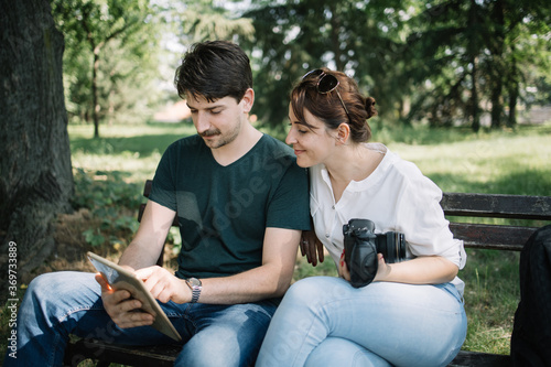 Woman and man sitting on bench and looking at tablet's display. Man and woman with camera checking photos on tablet while sitting on bench in park.
