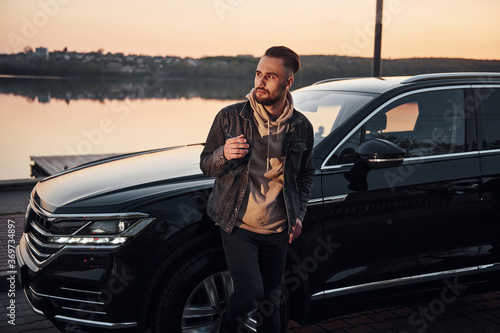 Handsome unshaved man in fashionable clothes standing near his black car and smoking © standret
