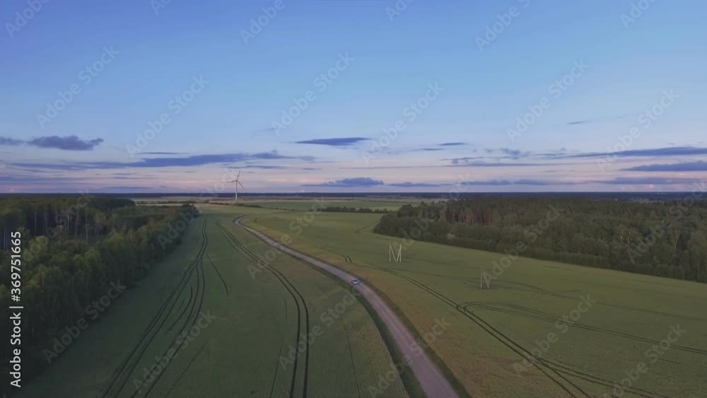 The Car Drives Through A Gravel Road Towards A Wind Turbine In Agricultural Fields. Aerial Drone Flight Forward The Car Drives Through A Gravel Road Towards A Wind Turbine In Agricultural Fields. Aerial Drone Flight Forward