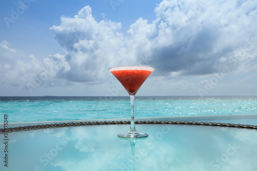 Glass of strawberry daiquiri with calm sea and clouds in Maldives sky.