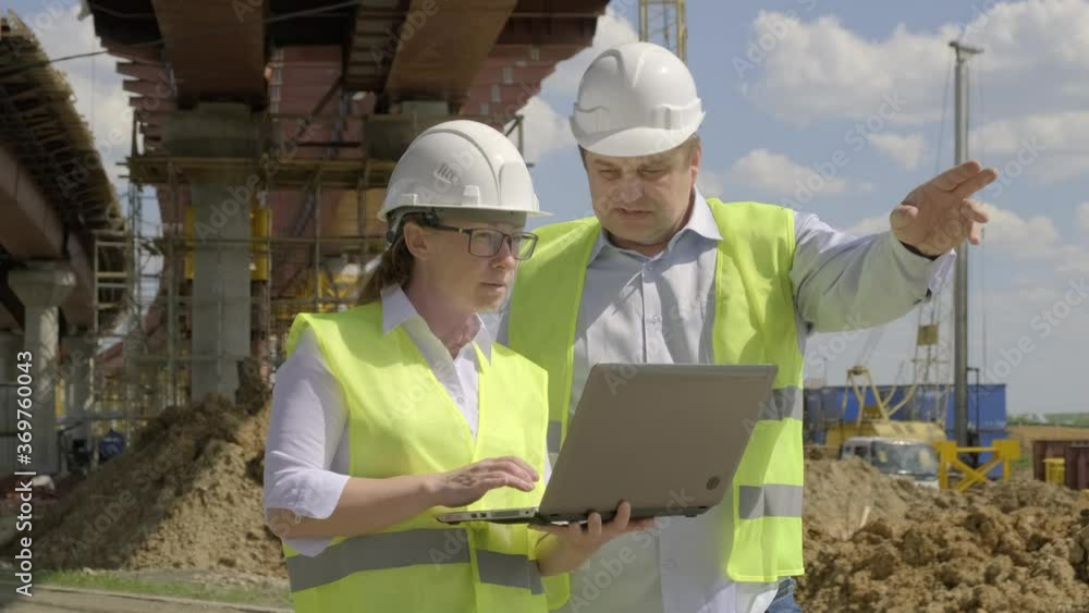 man and woman working on background of construction overpass. Builders discussing future work plan for erection of bridge using computer. man and woman working on background of construction overpass. Builders discussing future work plan for erection of bridge using computer.