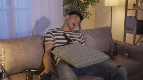 chinese guy playing game until wee hour fell asleep in messy living room out of fatigue. asian gamer napping with open mouth on couch scattered with bottles is out of energy. Carefree Holiday concept