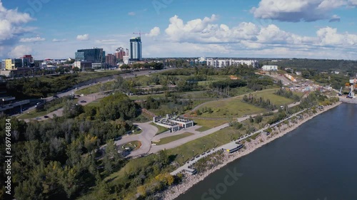 Aerial flyover the dark blue North Saskatchewan River, flyby the Louise Mckinney Public lush Park Chinese Garden Pavilion and oval track birds eye view on a hot summer day with cumulus cloud coverage photo