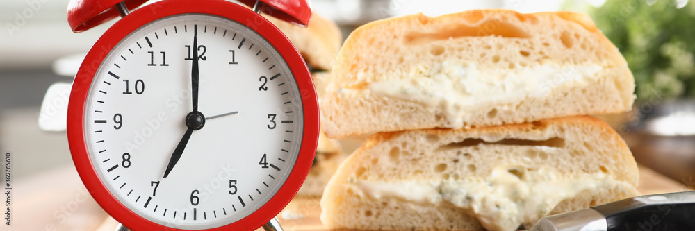 Table an alarm clock and breakfast bread and sauce. Sliced white bun with sauce. On table lies sandwich prepared at home during self-isolation. Delicious not healthy breakfast at home Table an alarm clock and breakfast bread and sauce. Sliced white bun with sauce. On table lies sandwich prepared at home during self-isolation. Delicious not healthy breakfast at home