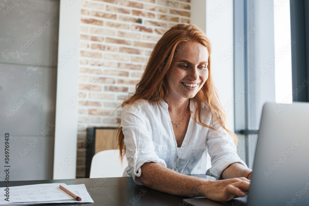 Image of smiling redhead businesswoman working with laptop while sitting Image of smiling redhead businesswoman working with laptop while sitting