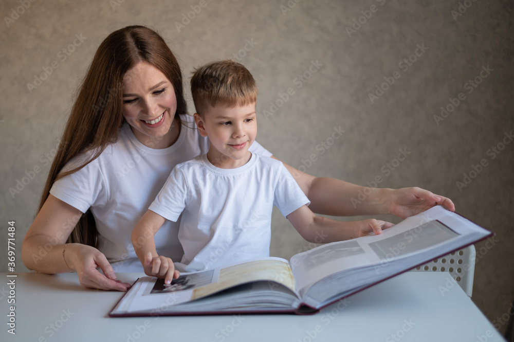 A middle-aged Caucasian woman hugs a little son and shows a photo album. A little boy sits on his mothers lap and looks at old photographs. Pleasant memories. Close family relationships. A middle-aged Caucasian woman hugs a little son and shows a photo album. A little boy sits on his mothers lap and looks at old photographs. Pleasant memories. Close family relationships.