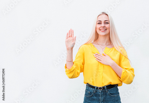 Attractive cute lovely blonde young girl raise one hand hold palm heart promise give oath pledge loyalty swear not tell lie standing happily friendly look camera, white background