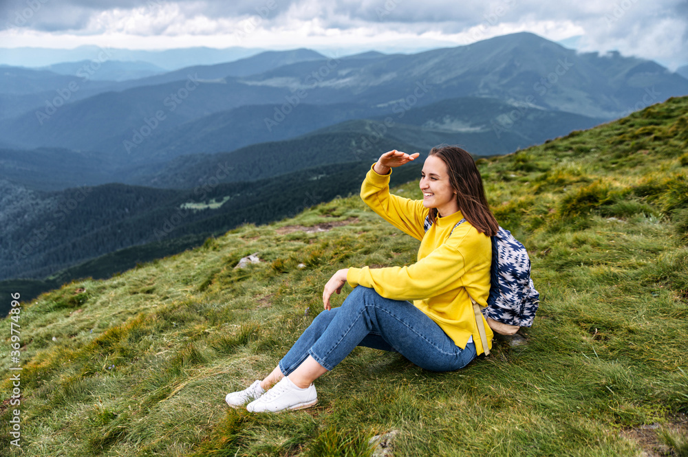 A beautiful tourist woman with a backpack rest after climbing to the top in summer mountains. A woman is looking into the distance with her hand near forehead to shield eyes from the sun as a visor