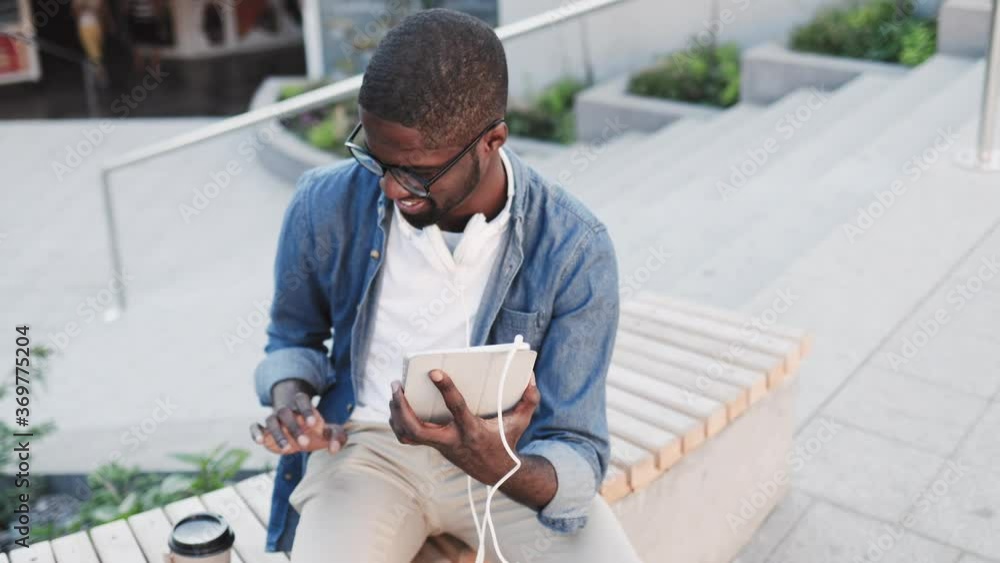 Happy African American man sitting on bench and using tablet. A guy with glasses sitting on street drinking coffee and playing on tablet. Happy African American man sitting on bench and using tablet. A guy with glasses sitting on street drinking coffee and playing on tablet.