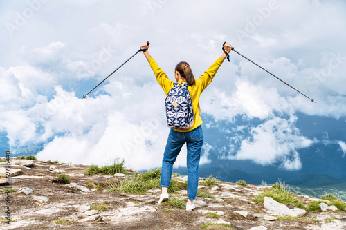 Female hiker enjoying a mountain view standing on the top. Back view a girl with a backpack put her hands up with a sticks for nordic walks