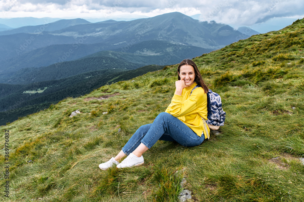 A beautiful young tourist woman with a backpack rest after climbing to the top in summer mountains. A woman is enjoying scenic landscape and relaxing