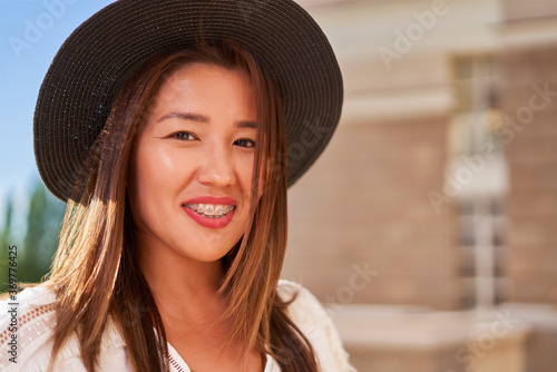 Braces on the teeth, a girl of Asian appearance in a white T-shirt and a dark hat, looking at the camera and smiling, blurred background