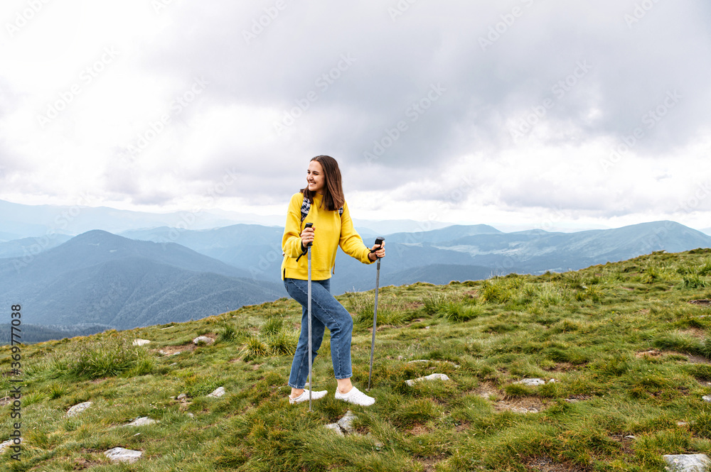 Naklejka premium A beautiful young tourist woman with a backpack is climbing to the top with a sticks for nordic walk in summer mountains. Active leisure time