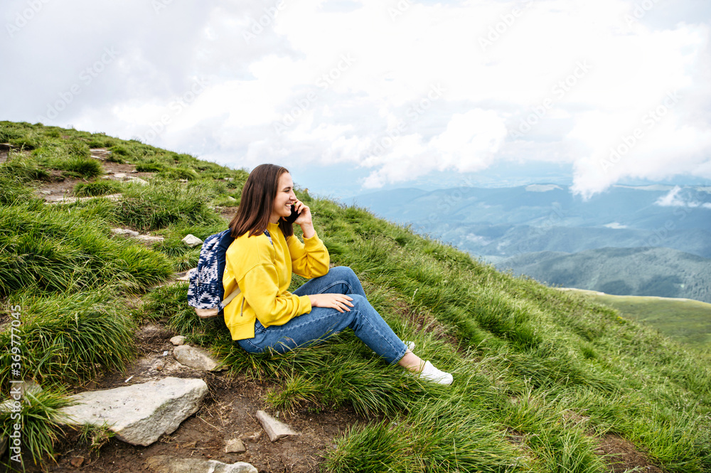 A hiker woman is talking on the smartphone while sitting on the top of the mountain. Backpacker girl speaks on the cellphone and smiles