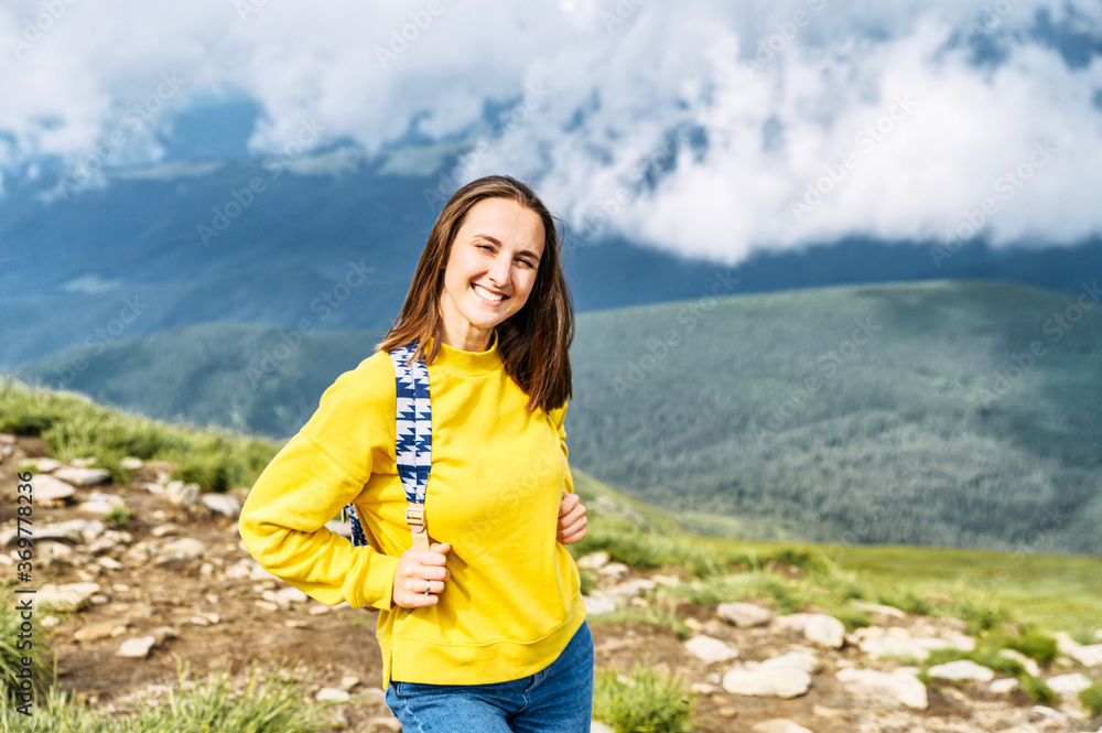 Naklejka premium Happy female hiker in the mountains. Cheerful girl with a backpack on the scenic nature and cloudy sky background