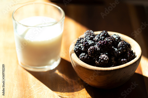 Large ripe Blackberries In a bowl and a glass of milk on a wooden background. Frosty berries are illuminated by bright sunlight. Selective focus. Minimalism. Close up