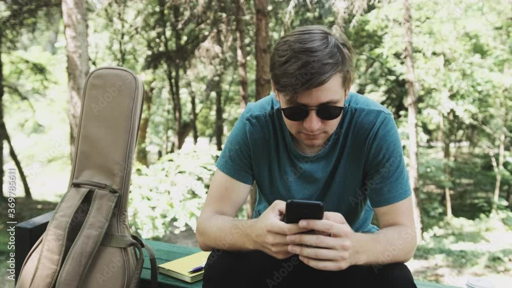 Young man with sun glasses is sitting on bench in park and watch the news in the internet. Young man with sun glasses is sitting on bench in park and watch the news in the internet.