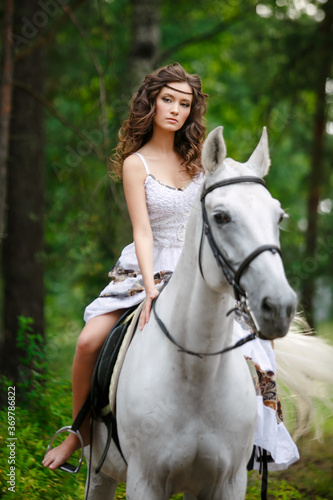 Young beautiful girl with white horse at summer green forest. Caucasian woman horseback rider in dress in boho style. Summertime scene