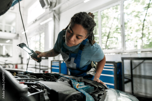 Moments of caring. Young african american woman, professional female mechanic looking, examining under hood of car with torch at auto repair shop © Svitlana