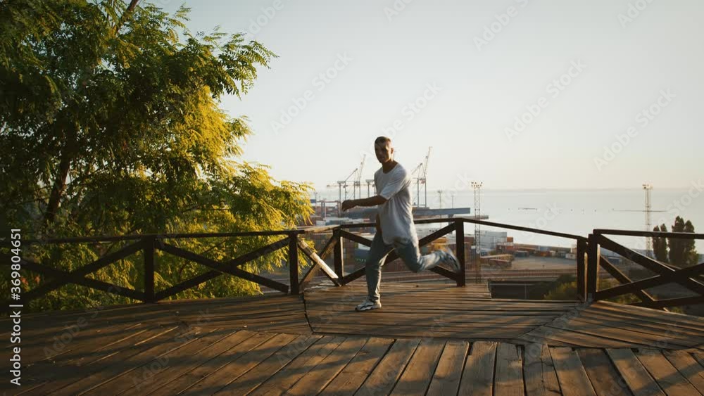 Young fellow is performing break dance on wooden terrace against picturesque seascape of city seaport. Morning, sunrise. Slow motion Young fellow is performing break dance on wooden terrace against picturesque seascape of city seaport. Morning, sunrise. Slow motion