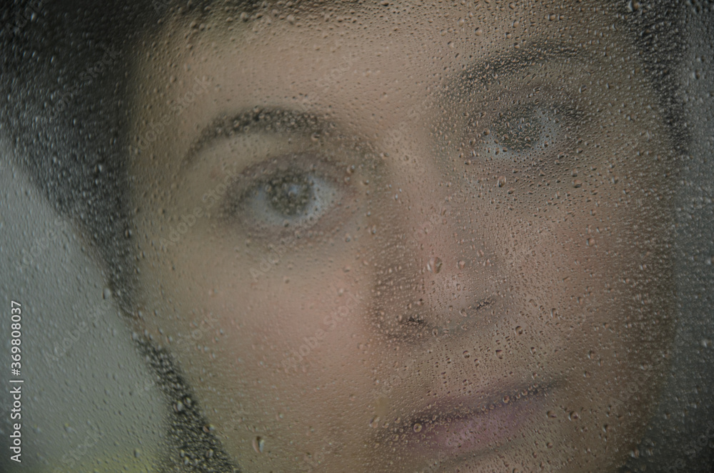 Portrait of a young brunette woman behind glass with rain drops. The concept of depression, blurring about the future, determination. Blur and selective focus on raindrops. Tinted image.
