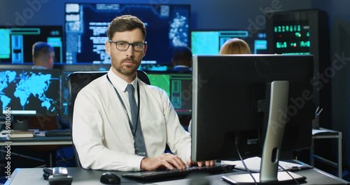 Portrait of Caucasian male official in glasses and tie working at computer and smiling to camera in analytic surveillance center. Handsome man checking info online in security anti-terrorism office. photo