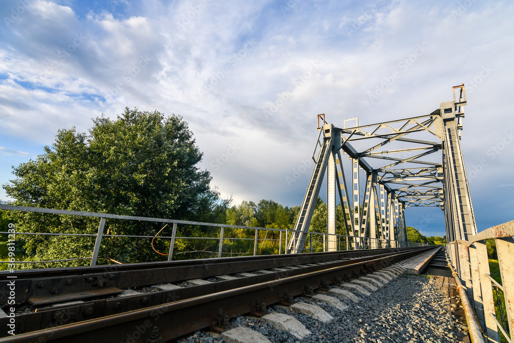 Fototapeta premium Railway bridge over the river at sunset in summer. Wide angle view. Background of blue sky.