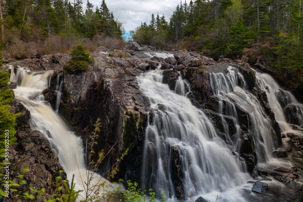 Multiple long natural waterfalls over a dark brown jagged rock cliff. The water is soft and white with some brown silt flowing over the edges. The background is blue sky, clouds and coniferous trees. Multiple long natural waterfalls over a dark brown jagged rock cliff. The water is soft and white with some brown silt flowing over the edges. The background is blue sky, clouds and coniferous trees.