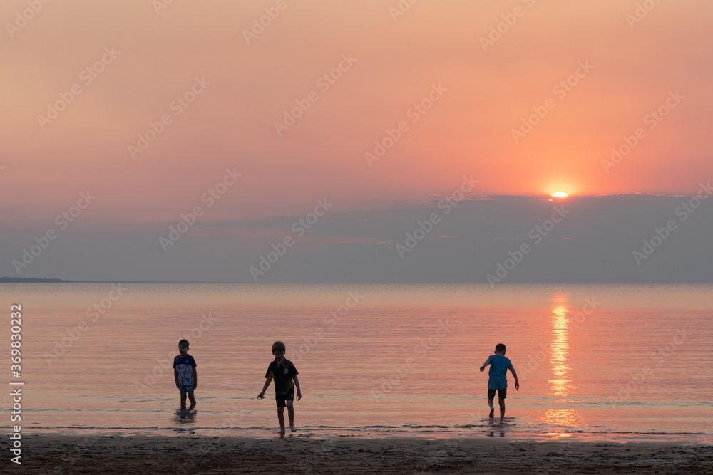 Kids playing at the beach at sunset time. Red and orange sky. Three young friends at the shore with feet in the ocean. Darwin, Northern Territory NT, Australia