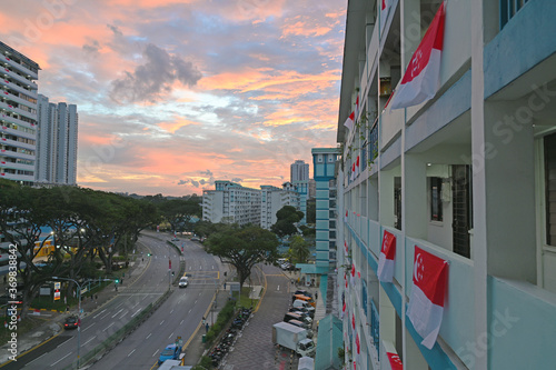 Singapore flags are hung outside some blocks, flutter against strong wind. The background is shined by light of sunset during month of August, which everyone celebrates Singapore's National Day.