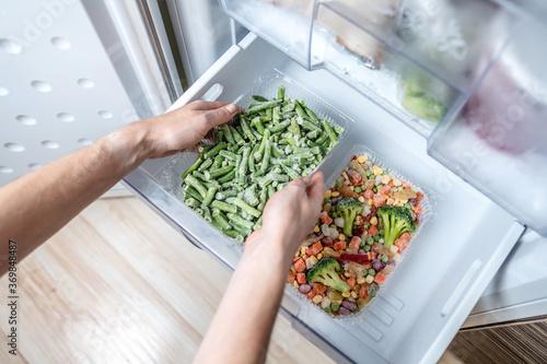 Hands are taking a container of frozen vegetables from the freezer of the fridge. Concept of storing ready dish and saving time photo