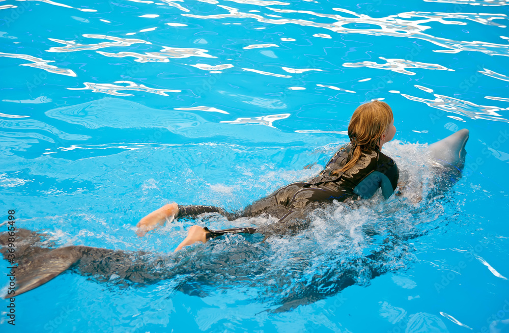 The girl in hydro-suit riding on a dolphin belly in blue water of dolphinarium