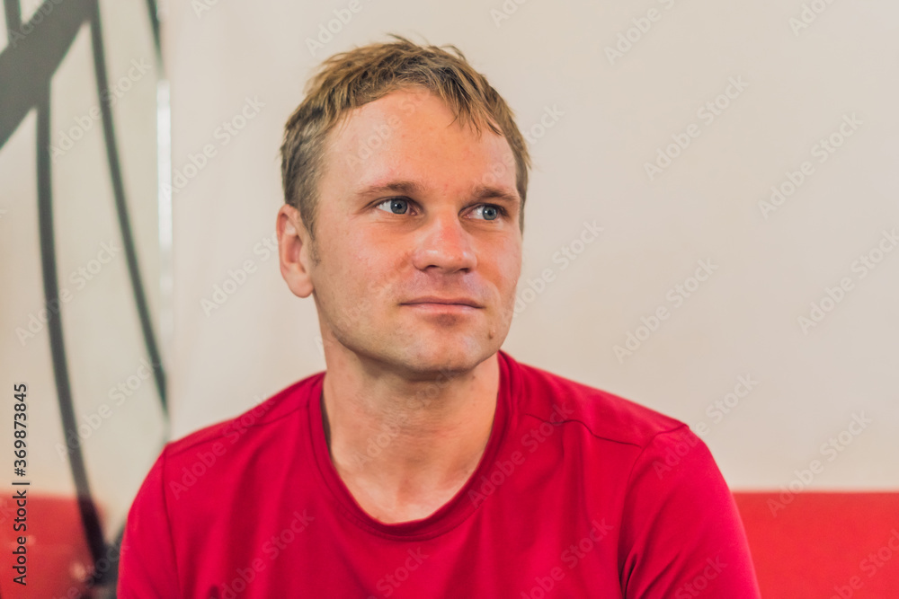 Front Portrait restrained smile sure handsome young blond man with mole in casual red t-shirt sit, look away, waiting thinking about changes good future or his girlfriend. Human relationship concept Front Portrait restrained smile sure handsome young blond man with mole in casual red t-shirt sit, look away, waiting thinking about changes good future or his girlfriend. Human relationship concept