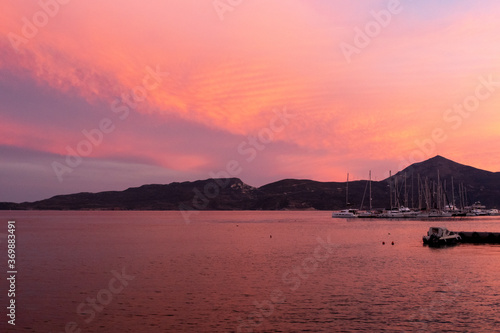 Fishing and sailing boats docked in Adamas Port on Milos Island, Greece during vivid pink sunset, with calm flat sea and pink cloudy sky, mountains in the background.
