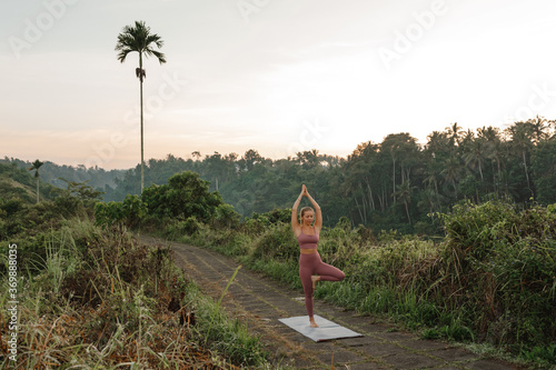 Young woman practicing tree yoga pose  in the park, beautiful tropical geenery landscape. Yoga retreat on Bali island photo