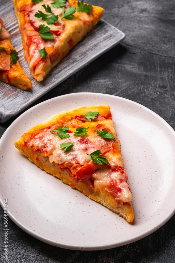 Hot pizza slices with mozzarella cheese, ham, tomato and parsley on wooden cutting board and plate, stone concrete background, angle view macro Hot pizza slices with mozzarella cheese, ham, tomato and parsley on wooden cutting board and plate, stone concrete background, angle view macro