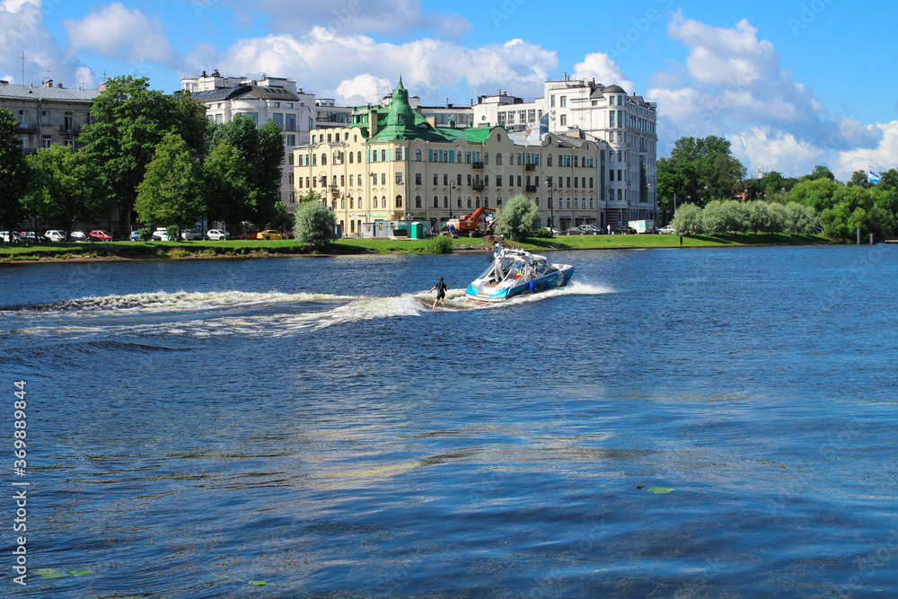 A trainer in a tracksuit with a megaphone from a boat leads a girl who starts wakesurfing against the background of the houses of St. Petersburg