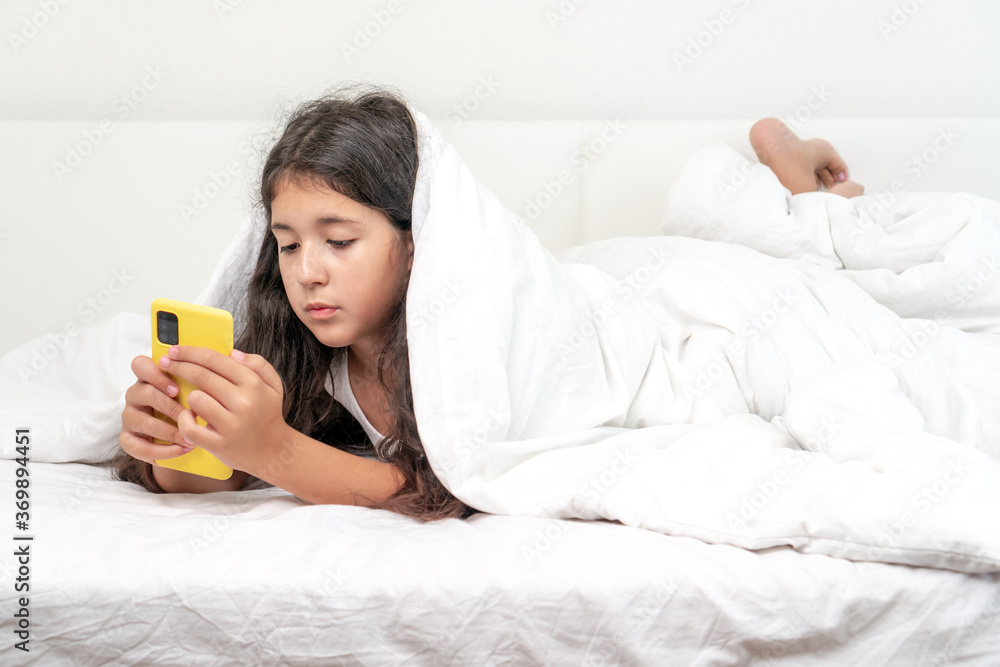 Cute little girl using a phone in bed under a blanket. A girl is watching their social networks on a phone. Remote education concept. Cute little girl using a phone in bed under a blanket. A girl is watching their social networks on a phone. Remote education concept.