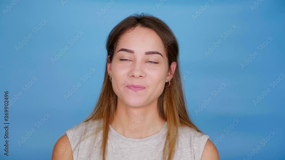 Beautiful funny woman in gray t-shirt talking, squinting and smiling on blue background with copy space. 4K slow motion footage Beautiful funny woman in gray t-shirt talking, squinting and smiling on blue background with copy space. 4K slow motion footage