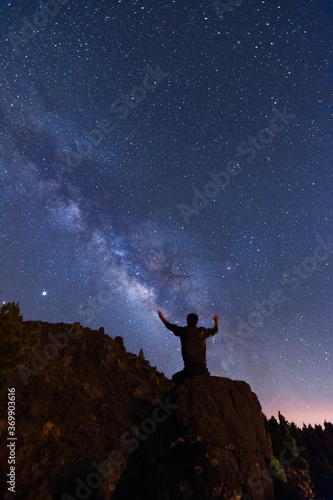 Young man with raised arms looking at the stars and the milky way on a summer night in the mountains
