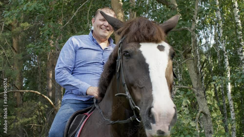Smiling horseman sitting astride horse on green trees background on farm ranch. Happy stableman sitting on horseback while horse walk in summer forest