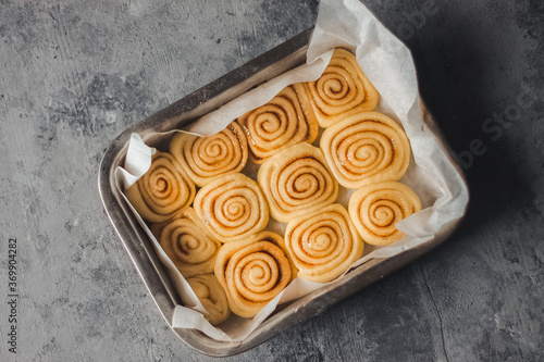 Delicious homemade raw cinnamon rolls bun dough in the rectangle shaped baking tray on the white baking paper. Top view.