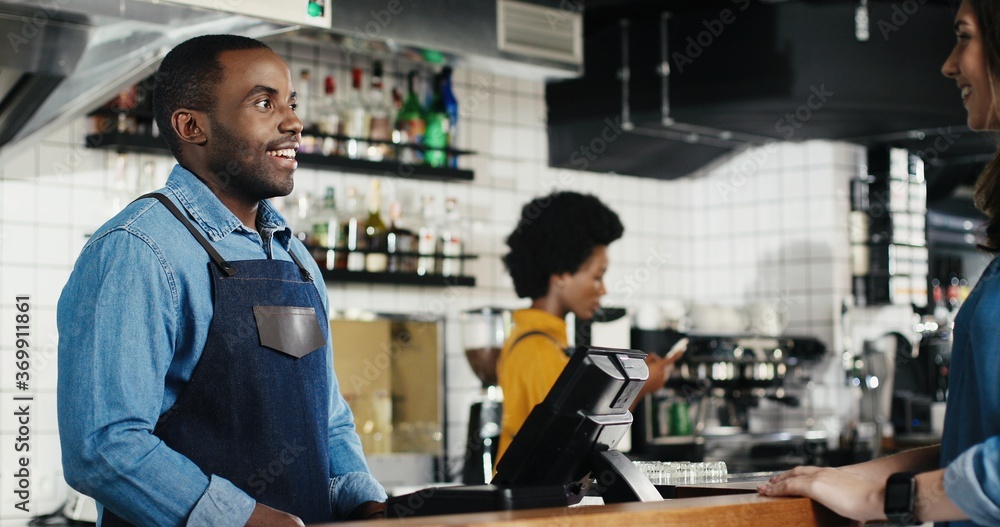 Cheerful African American waiter serving coffee and talking with Caucasian woman at counter in cafe. Man barrista having nice talk with customer. Male bartender chatting with female client Multiethnic Cheerful African American waiter serving coffee and talking with Caucasian woman at counter in cafe. Man barrista having nice talk with customer. Male bartender chatting with female client Multiethnic