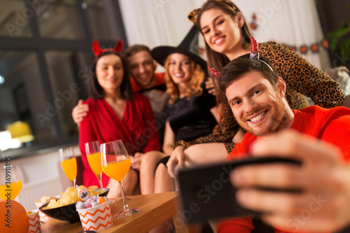 friendship  holiday and people concept - group of happy smiling friends in halloween costumes of vampire  devil  witch and cheetah taking selfie by smartphone at home party at night