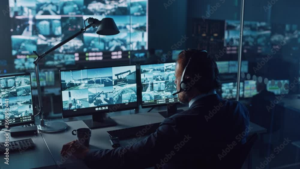 Male Officer Works on a Computer with Surveillance CCTV Video Footage in a Harbour Monitoring Center with Multiple Cameras on a Big Digital Screen. Employees Sit in Front of Displays with Big Data. Male Officer Works on a Computer with Surveillance CCTV Video Footage in a Harbour Monitoring Center with Multiple Cameras on a Big Digital Screen. Employees Sit in Front of Displays with Big Data.