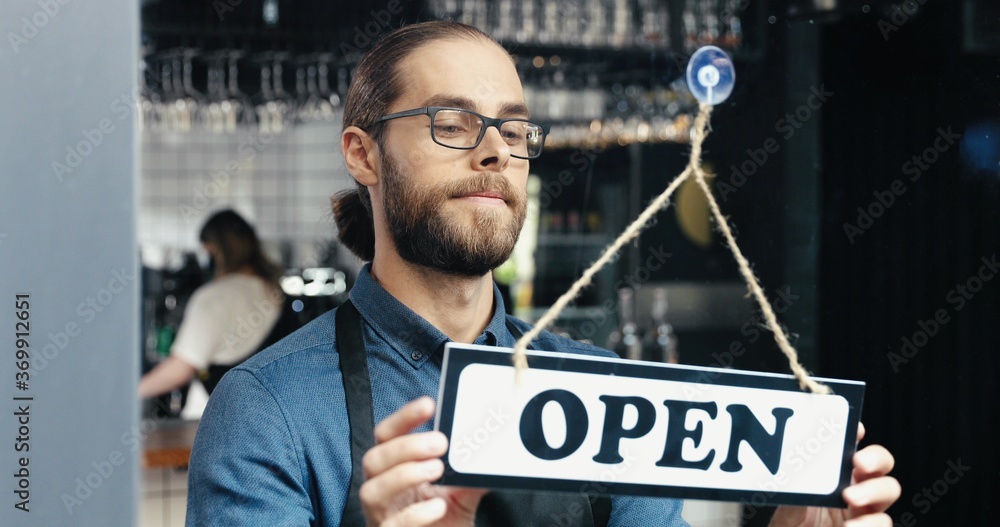 Close up of Caucasian handsome happy man waiter in glasses changing table on door - closed-open at cafe in the morning. Male opening bar early and smiling. Cheerful vendor turning board and opens shop