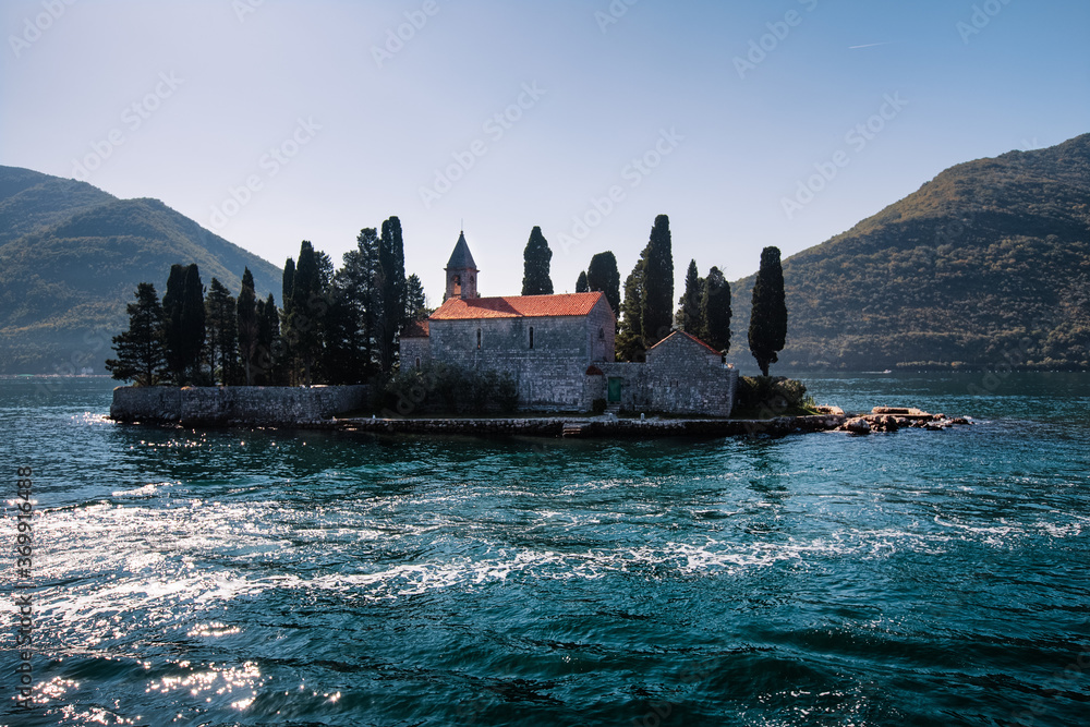 Catholic red roof medieval monastery, bell tower, masonry wall and cypress trees on an island in the Adriatic Sea. Little island in blue water of Kotor bay among Montenegro mountains. Island panorama