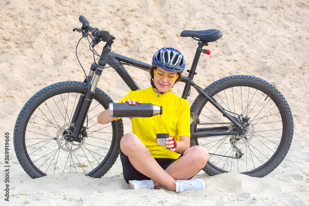 Naklejka premium Beautiful girl cyclist pours tea from a thermos on the background of a Bicycle in the sand. Sports and recreation. Hobbies and health.